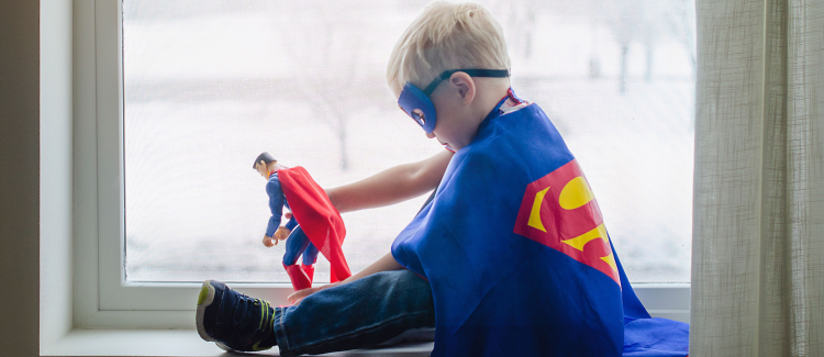 child sitting in window wearing blue superhero cape, playing with a supehero