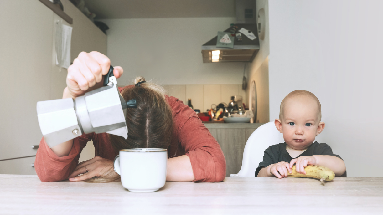 tired mom pouring coffee with baby eating a banana beside her.
