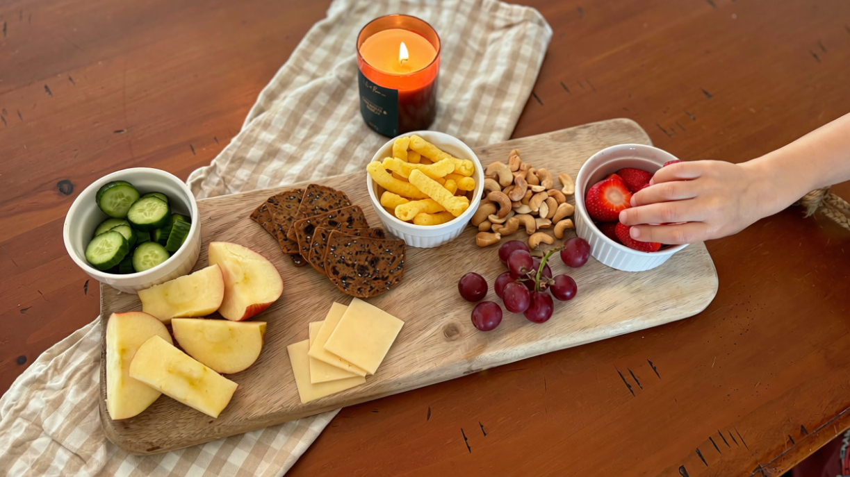 a photo of snacks on a board and a child's hand reaching for a strawberry.